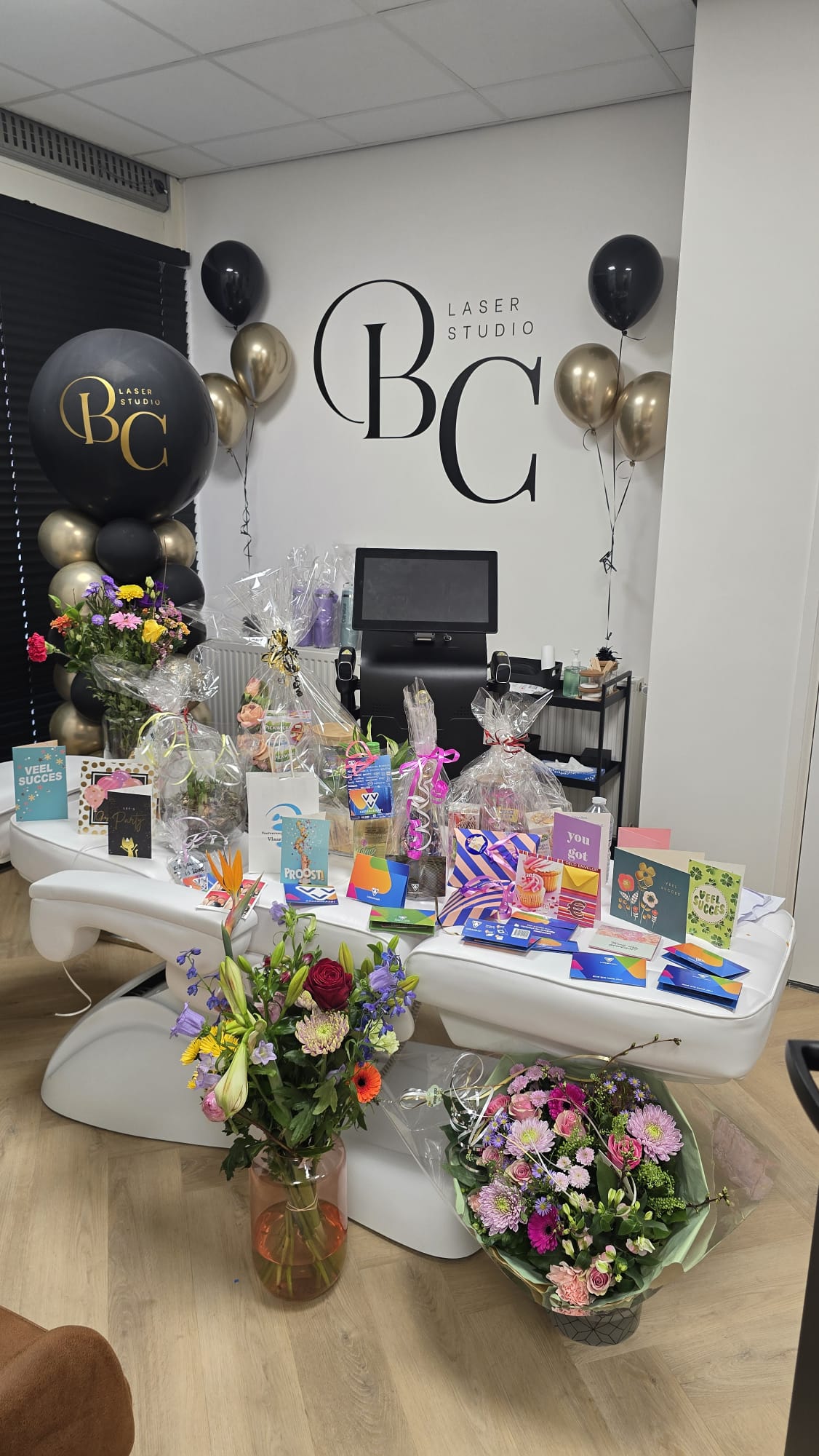Bloemen, kaarten en zwarte en gouden ballonnen onder het BC-logo op de muur, bij de opening